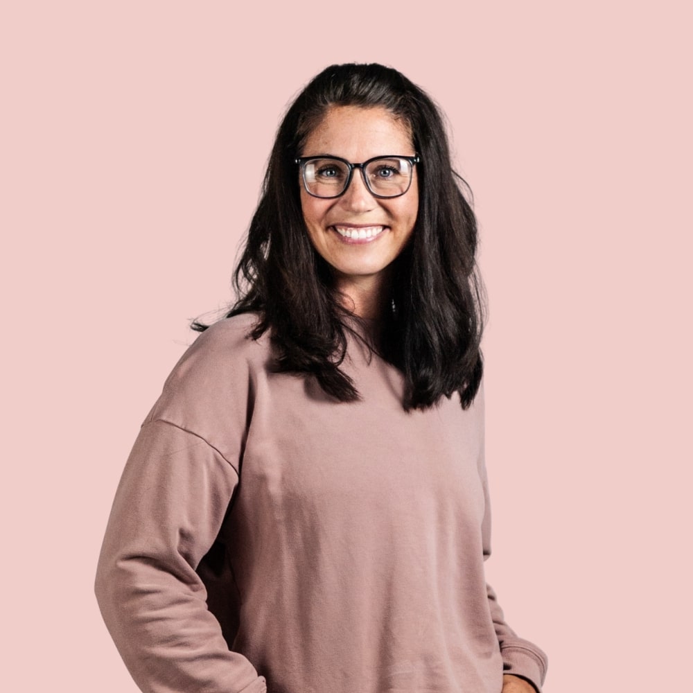 Smiling person with long dark hair and glasses wearing a mauve sweater stands against a plain pink background.