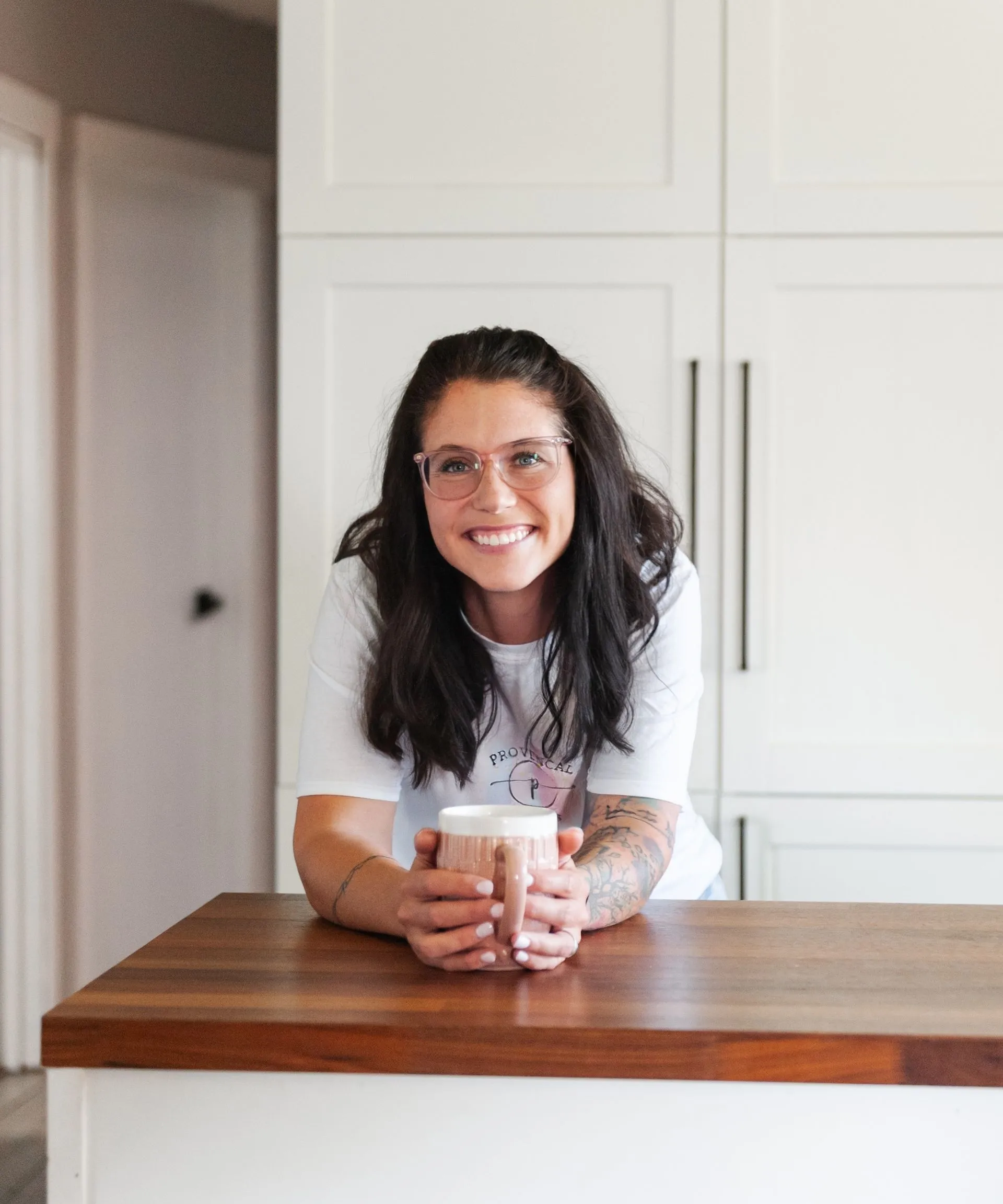 Smiling woman with glasses and long dark hair holds a mug, leaning on a wooden countertop in a kitchen with white cabinets.