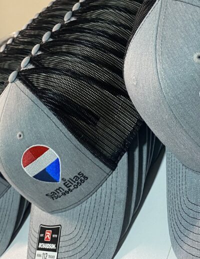 Row of gray and black trucker hats with a red, white, and blue logo, featuring "sam elias" and phone number embroidery on the front.