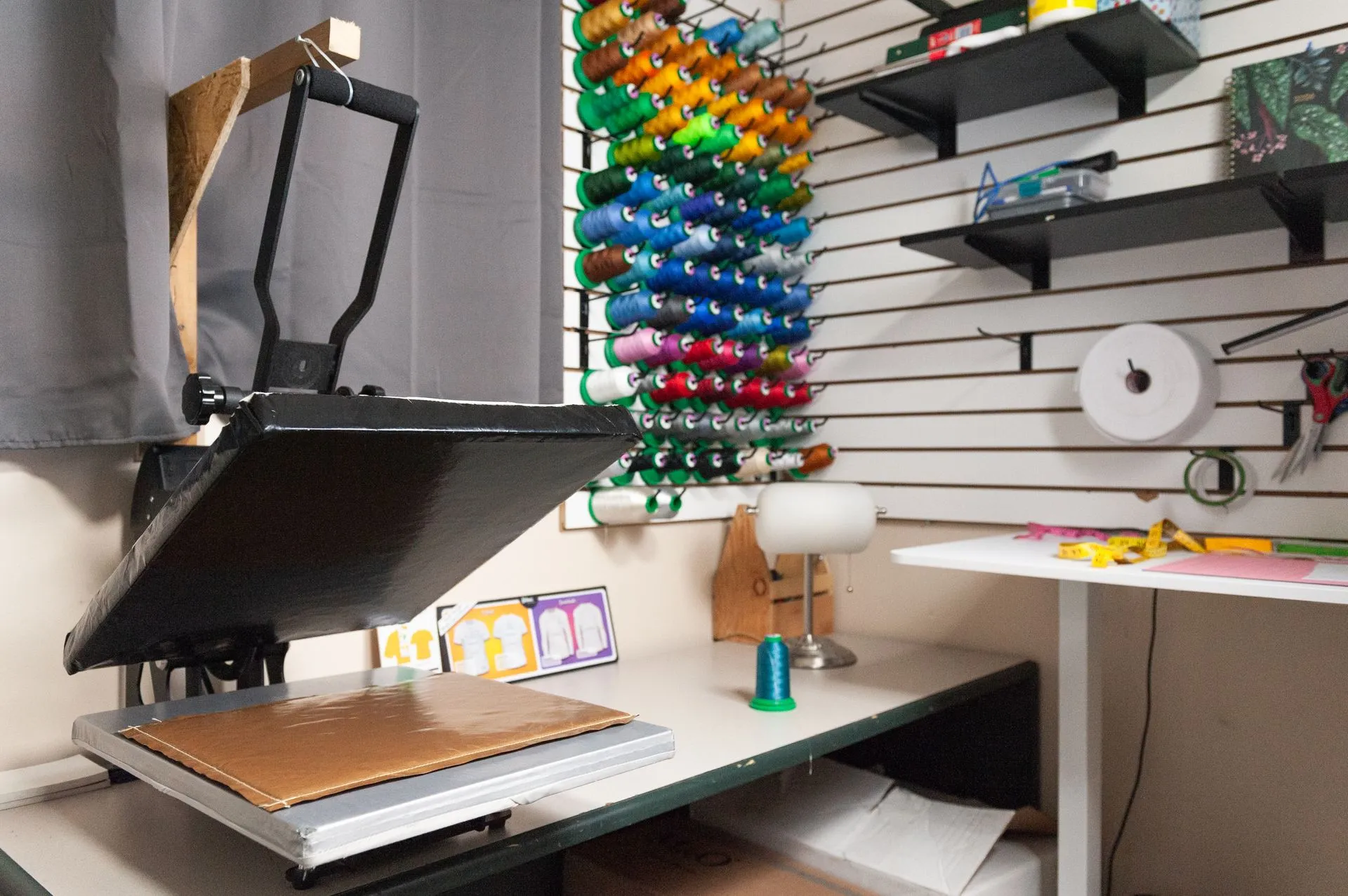 Heat press machine in a workspace with colorful thread spools on the wall, shelves with supplies, and a table with tools like scissors and