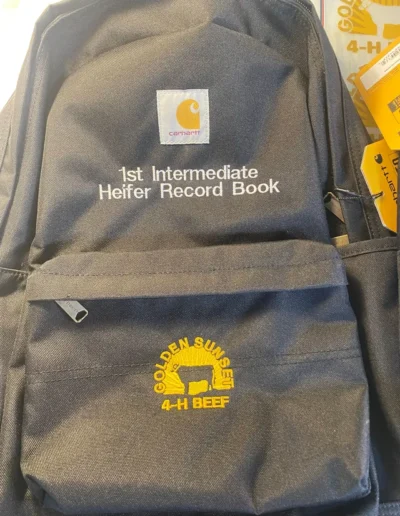 Dark blue backpack with white "1st Intermediate Heifer Record Book" text and "Golden Sunset 4-H Beef" yellow logo featuring a cow silhouette.