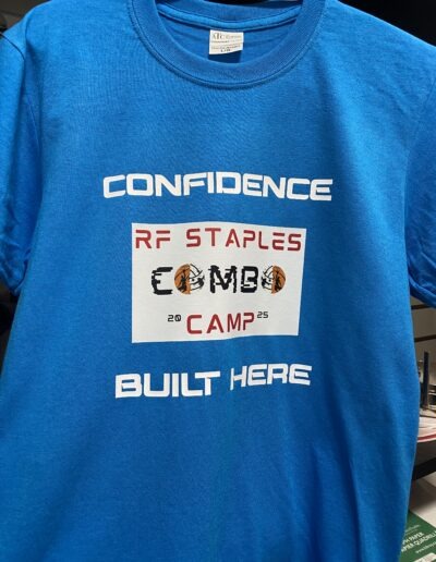 Blue T-shirt with bold white text "CONFIDENCE BUILT HERE," featuring a logo for "RF Staples Combo Camp 2025" with basketball graphics.