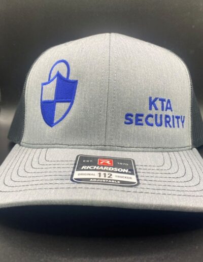 Gray trucker hat with blue embroidered shield and "KTA Security" text. Front features a "Richardson 112" label. Black mesh back, displayed in a